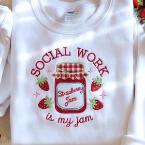 May include: White sweatshirt with an embroidered design featuring a strawberry jam jar and strawberries. The text reads "SOCIAL WORK is my jam" in red script. The jar has a red and white checkered lid.