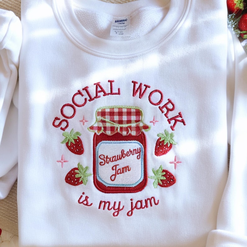 May include: White sweatshirt with an embroidered design featuring a strawberry jam jar and strawberries. The text reads "SOCIAL WORK is my jam" in red script. The jar has a red and white checkered lid.