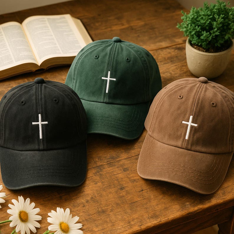 May include: Three baseball caps in black, green, and brown, each with a white cross embroidered on the front. The caps are displayed on a wooden surface, with a book and a small potted plant in the background.