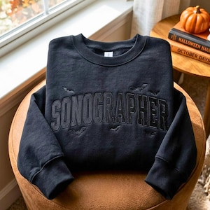 May include: A black crewneck sweatshirt featuring the word SONOGRAPHER embroidered in a dark, arched collegiate font across the chest. The design includes small, subtle embroidered bat silhouettes scattered around the text on the front.