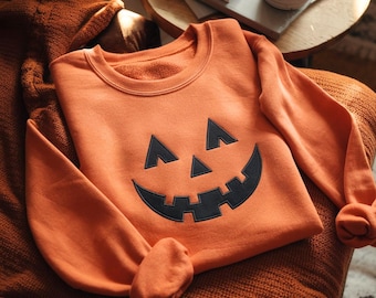 Op de afbeelding: Oranje sweatshirt met een zwart pompoengezicht. Het ontwerp omvat driehoekige ogen en een brede, tandige grijns. Dit zachte sweatshirt is ideaal voor Halloween. Gemaakt van een comfortabel materiaal.