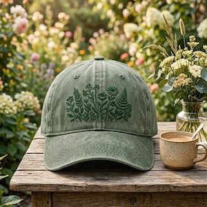 May include: A green baseball cap with an embroidered floral design. The cap is made of a soft, worn fabric and features a curved brim. A cup of coffee and a vase of flowers are on the table.