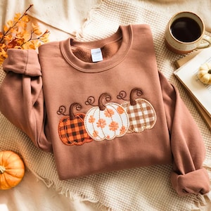 May include: A rust-colored crewneck sweatshirt featuring an embroidered design of three pumpkins across the chest. The left pumpkin has a black and orange buffalo check pattern, the center is white with orange floral details, and the right is brown gingham.