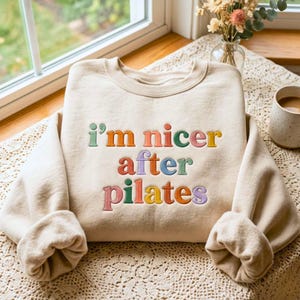 Puede incluir: Una sudadera de cuello redondo en color beige con el texto bordado "i'm nicer after pilates" en una fuente multicolor. Las letras están cosidas en tonos verde, naranja, azul, amarillo y morado en el centro de la zona del pecho.