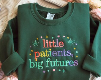 Puede incluir: Una sudadera verde oscuro con la frase bordada "little patients, big futures" en letras coloridas. El diseño incluye estrellas y pequeños detalles florales. La sudadera tiene mangas largas y cuello redondo.