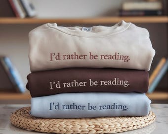 May include: A stack of three sweatshirts in cream, brown, and light blue, each embroidered with the phrase "I'd rather be reading." The sweatshirts are neatly folded and stacked on a woven placemat, suggesting a cozy, book-themed aesthetic.