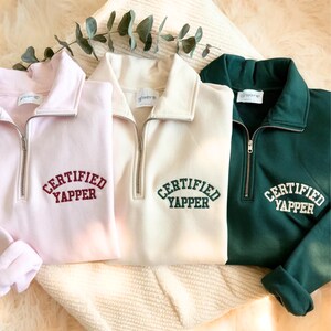 May include: Three quarter-zip sweatshirts in pink, cream, and green. Each sweatshirt has the embroidered text "CERTIFIED YAPPER" across the chest. The sweatshirts have a collared neckline and a silver zip.