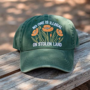May include: Green baseball cap with the text "NO ONE IS ILLEGAL ON STOLEN LAND" and a floral design in orange and white. The cap is sitting on a wooden surface.