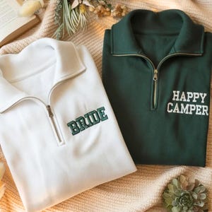May include: Two zip-up sweatshirts, one white and one dark green, are displayed. The white sweatshirt has "BRIDE" embroidered in green, while the green one says "HAPPY CAMPER" in white. Both have a collared neckline and a zipper.