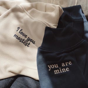 K&ouml;nnte beinhalten: Zwei Sweatshirts, eines in Creme und eines in Marineblau, werden gezeigt. Das cremefarbene Sweatshirt hat den schwarzen Schriftzug "I love you mostest". Das marineblaue Sweatshirt hat den wei&szlig;en Schriftzug "you are mine".
