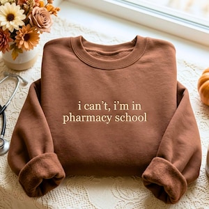 May include: A brown crewneck sweatshirt featuring the text "i can't, i'm in pharmacy school" printed in a light beige font across the center. The garment has long sleeves with ribbed cuffs and a ribbed neckline, showcasing a simple design.
