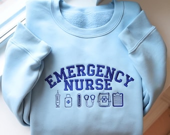 May include: A light blue sweatshirt with the words "EMERGENCY NURSE" in navy blue lettering. Below the text are navy blue medical-themed icons, including a syringe, a medicine bottle, a thermometer, a stethoscope, a medical bag, and a clipboard.