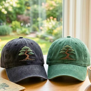May include: Two baseball caps, one in charcoal gray and one in sage green, each featuring an embroidered design of a bonsai tree with a brown trunk and green foliage centered on the front panel. Both caps have a curved brim and a structured crown.
