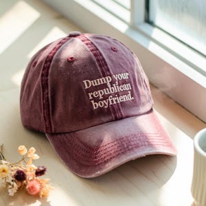 May include: A maroon baseball cap with the text "Dump your republican boyfriend" embroidered in white. The cap is made of a soft, worn material and has a curved brim. The cap is sitting on a wooden surface.