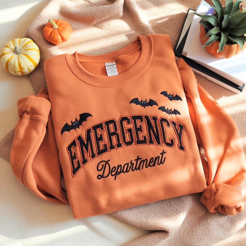 May include: An orange sweatshirt with the words "EMERGENCY Department" in black lettering and bat graphics. The sweatshirt is displayed on a soft, textured surface, with small pumpkins and a book nearby. A cozy, seasonal theme.