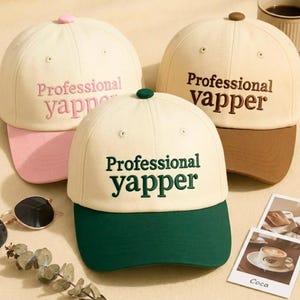 May include: Three baseball caps with cream-colored crowns and contrasting brims. One cap has a pink brim and pink embroidered text reading Professional yapper. Another has a brown brim and brown text reading Professional vapper. The third has a green brim and green text reading Professional yapper.