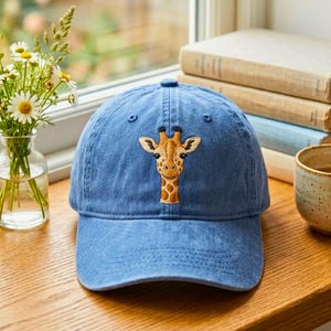 Puede incluir: Una gorra de béisbol azul con una cabeza de jirafa bordada en el panel frontal. La gorra tiene una visera curva, un botón en la parte superior y ojales cosidos para la ventilación. El diseño de la jirafa es en tonos marrón y beige.