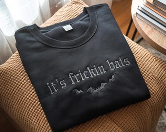 May include: Black sweatshirt with the embroidered phrase "it's frickin bats" in a gothic font, along with bat silhouettes. The sweatshirt is displayed on a brown cushion, with a small table and books in the background.