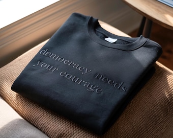 May include: A black sweatshirt with the embossed text "democracy needs your courage." The crew neck sweatshirt is folded and resting on a brown cushion. The image is taken in natural light, with a wooden table and books in the background.