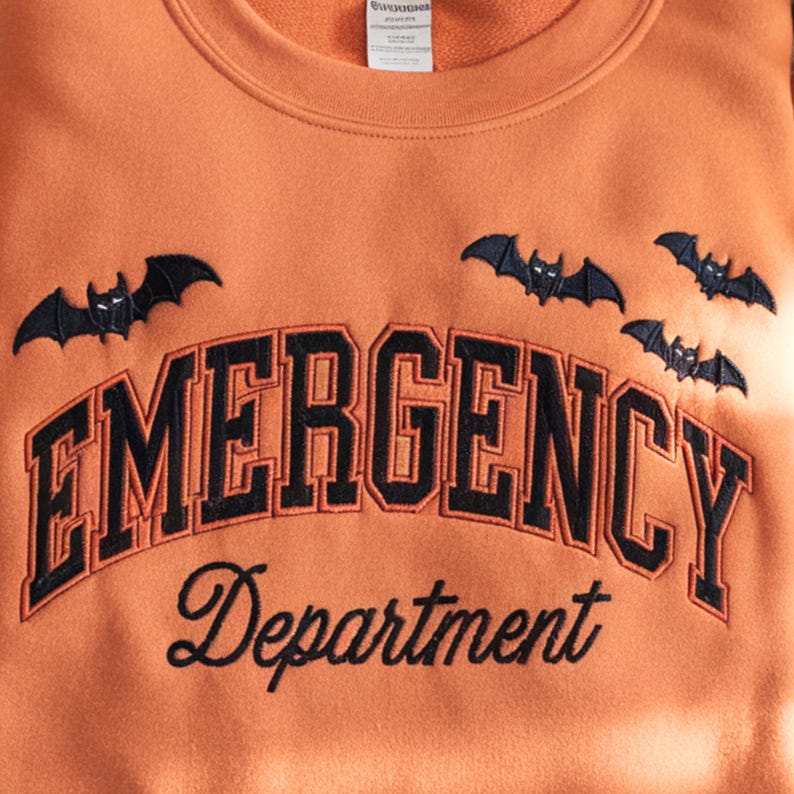 May include: Orange sweatshirt with the words "EMERGENCY Department" in black and navy blue embroidery. Above the text are three embroidered black bats. The sweatshirt has a crew neck.