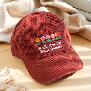 Könnte beinhalten: Eine ziegelrote Cord-Baseballkappe mit gestickten Blumen und dem Text "Radicalized by Basic Decency". Die Kappe hat einen gebogenen Schirm und eine strukturierte Krone. Die Blumen sind in Gelb-, Rosa- und Rottönen gehalten.