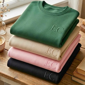 May include: A stack of four crewneck sweatshirts in forest green, beige, light pink, and black. Each sweatshirt features the embroidered letters EC on the upper right chest area. The garments are folded neatly and stacked vertically on a wooden surface.