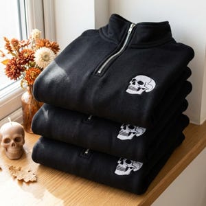 May include: A stack of three black quarter-zip sweatshirts, each featuring a white embroidered skull design on the chest. The sweatshirts have a high collar with a silver-toned metal zipper closure, showcasing a consistent design across all items.