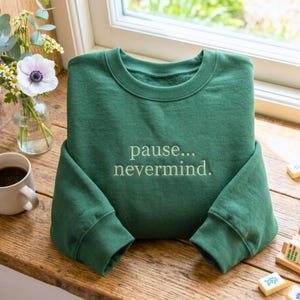 May include: A forest green crewneck sweatshirt featuring the embroidered text "pause... nevermind." in a lighter green thread. The garment has long sleeves with ribbed cuffs and a ribbed neckline, presented in a folded position on a wooden surface.