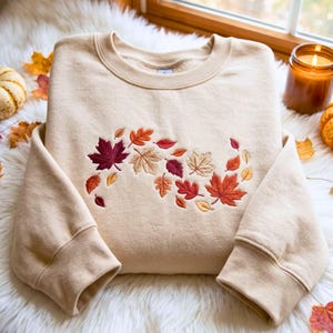 May include: A beige crewneck sweatshirt featuring an embroidered design of scattered autumn leaves in shades of deep red, orange, gold, and brown across the chest. The garment has long sleeves with ribbed cuffs and a ribbed neckline.