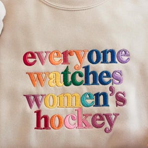 K&ouml;nnte beinhalten: Beiges Sweatshirt mit dem gestickten Text "everyone watches women's hockey" in Regenbogenfarben. Der Text ist in einer fetten, verspielten Schriftart gehalten, ein Statement f&uuml;r Frauensport.