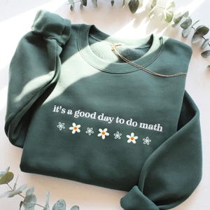 May include: A dark green sweatshirt with the phrase "it's a good day to do math" in white script. Below the text are six white daisies with orange centers. A gold necklace rests on the sweatshirt.