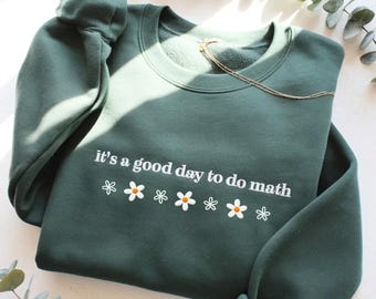 May include: A dark green sweatshirt with the phrase "it's a good day to do math" in white script. Below the text are six white daisies with orange centers. A gold necklace rests on the sweatshirt.