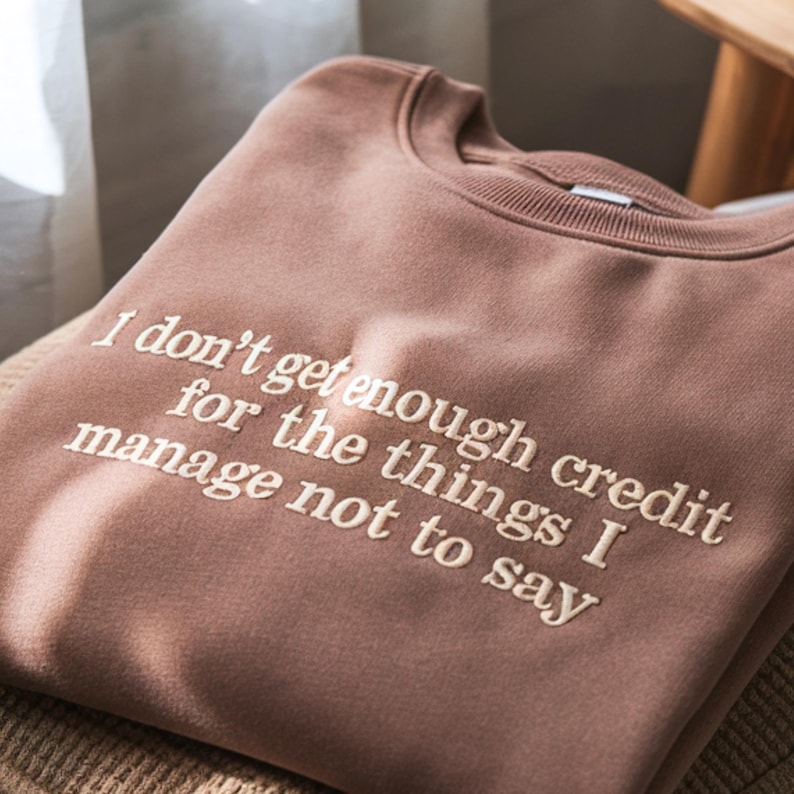 May include: A brown sweatshirt with the embroidered text "I don't get enough credit for the things I manage not to say." The crew neck sweatshirt is folded on a patterned surface.
