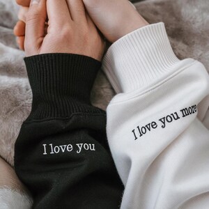 K&ouml;nnte beinhalten: Nahaufnahme von zwei passenden Sweatshirts, deren &Auml;rmel sich ber&uuml;hren. Ein &Auml;rmel ist schwarz mit dem gestickten Text "I love you", und der andere ist wei&szlig; mit dem Text "I love you more". Die B&uuml;ndchen sind gerippt.