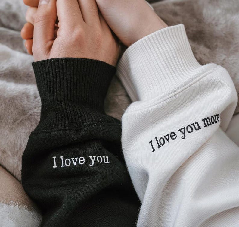 K&ouml;nnte beinhalten: Nahaufnahme von zwei passenden Sweatshirts, deren &Auml;rmel sich ber&uuml;hren. Ein &Auml;rmel ist schwarz mit dem gestickten Text "I love you", und der andere ist wei&szlig; mit dem Text "I love you more". Die B&uuml;ndchen sind gerippt.