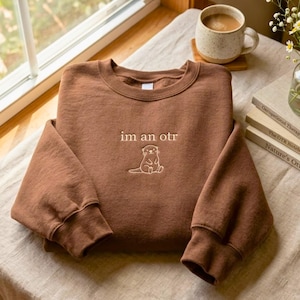 May include: A brown sweatshirt with the embroidered text "im an otr" and a small otter graphic. The long-sleeved crewneck top is folded on a neutral surface, with a coffee mug and books in the background.