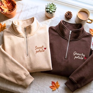 May include: A beige quarter-zip sweatshirt with the embroidered text Sweet potato and a small red heart, and a dark brown quarter-zip sweatshirt with the embroidered text Grouchy potato and a small red heart, both featuring silver zippers.