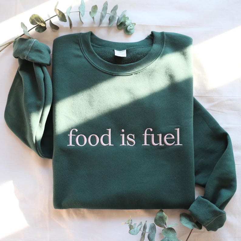 May include: A dark green sweatshirt with the embroidered text "food is fuel" in white. The sweatshirt has a crew neck and long sleeves, and is displayed on a white surface with greenery.