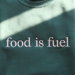 May include: A dark green sweatshirt featuring the embroidered words "food is fuel" in pale pink. The casual crew neck top is made of a soft fabric. The text is centrally placed on the chest, suitable for everyday wear.