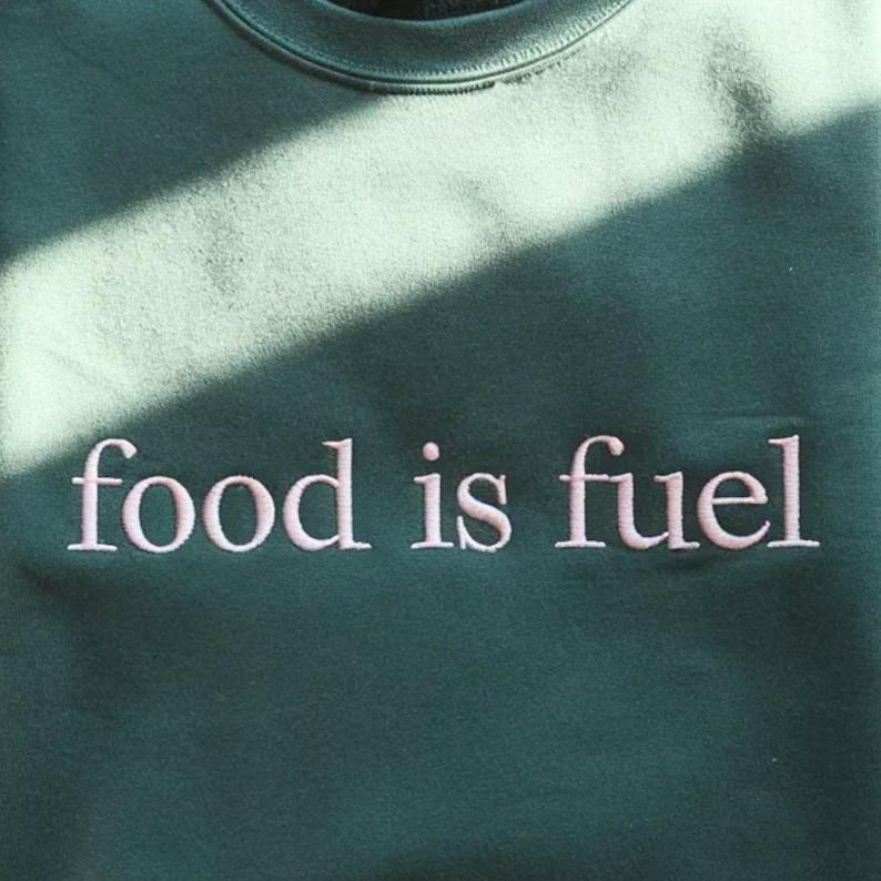 May include: A dark green sweatshirt featuring the embroidered words "food is fuel" in pale pink. The casual crew neck top is made of a soft fabric. The text is centrally placed on the chest, suitable for everyday wear.