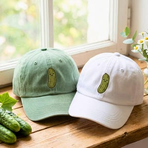 May include: Two baseball caps, one in sage green and one in white, each featuring an embroidered green pickle character with a smiling face on the front panel. The caps have a structured crown, a curved brim, and matching top buttons.