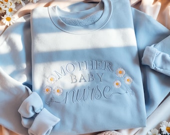 May include: A light blue sweatshirt with the embroidered text "MOTHER BABY nurse" in a cursive font, adorned with small daisy flower details. The sweatshirt has a crew neck and long sleeves.