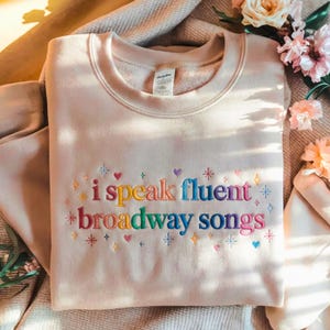 May include: A beige sweatshirt with the embroidered text "i speak fluent broadway songs" in rainbow colors. The design includes hearts, stars, and diamonds. The sweatshirt is folded on a soft, neutral-colored surface.