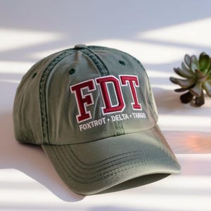 Könnte beinhalten: Olivgrüne Baseballkappe mit den Buchstaben "FDT" in Burgunderrot und Weiß, darunter die Worte "Foxtrot Delta Tango". Die Kappe besteht aus einem weichen Material und hat einen gebogenen Schirm. Ein lässiges Accessoire.