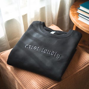 May include: Black sweatshirt with the phrase "exist loudly" embroidered in a gothic font. The sweatshirt is folded neatly on a corduroy cushion. The background includes a wooden side table and a window with natural light.