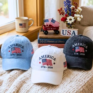 May include: Three baseball caps in light blue, white, and dark gray, each embroidered with "AMERICA", a waving American flag, and "1776-2026". The caps have a curved brim and a small American flag on the side. A patriotic theme.
