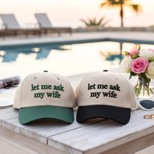 May include: Two baseball caps with the text "let me ask my wife". One cap has a green brim, the other has a black brim. Both caps are beige with black text. The caps are on a white wooden table near a pool.