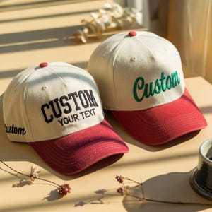 May include: Two baseball caps with off-white crowns and red brims. The left cap features black embroidered text reading CUSTOM YOUR TEXT and custom on the side. The right cap features green embroidered text reading Custom on the front.