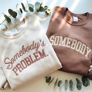 Puede incluir: Se muestran dos sudaderas de cuello redondo, una beige y otra marrón. La sudadera beige tiene el texto bordado "Somebody's PROBLEM." en marrón rojizo. La sudadera marrón tiene el texto bordado "SOMEBODY" en crema.