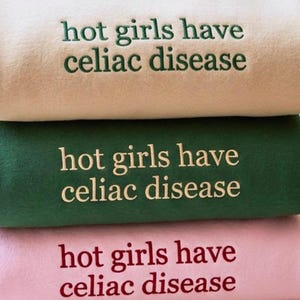 May include: A stack of three folded sweatshirts in beige, dark green, and light pink. Each sweatshirt features the embroidered text "hot girls have celiac disease" in a contrasting color: dark green on beige, beige on green, and dark red on pink.